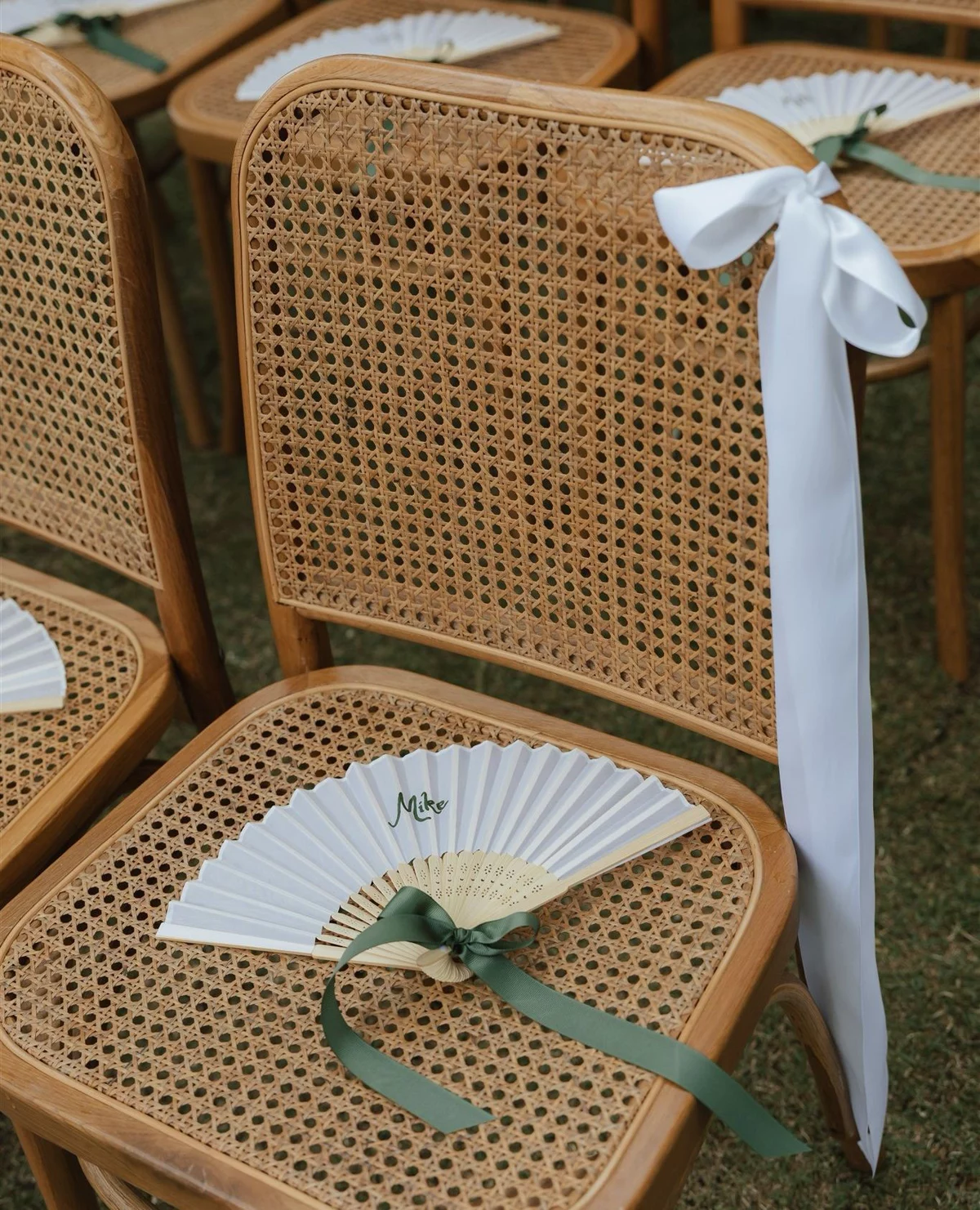 White fan wedding favors with sage ribbon placed on rattan ceremony chairs